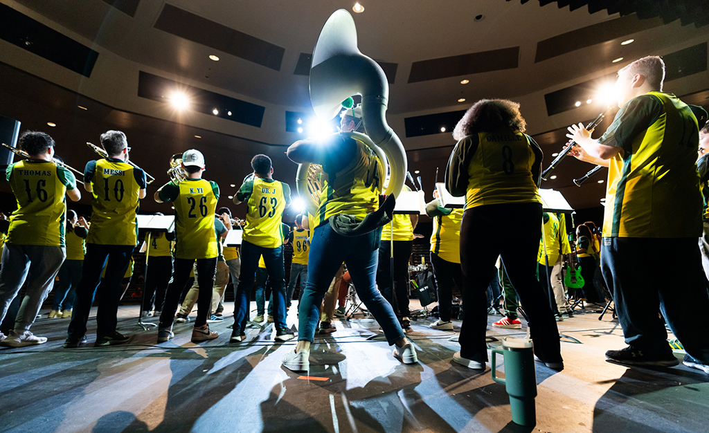 Members of the green machine perform indoors in an auditorium. The photographer is behind the band looking up from the floor through the band into the audience. The band is dancing and playing and is wearing yellow jerseys.