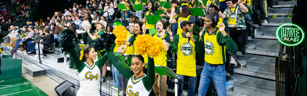 The Green Machine Pep Band performs in the stands in EagleBank Arena during a basketball game. The band is cheering along with cheerleaders in white uniforms. The band is in yellow basketball jerseys. The arena is full of fans screaming.