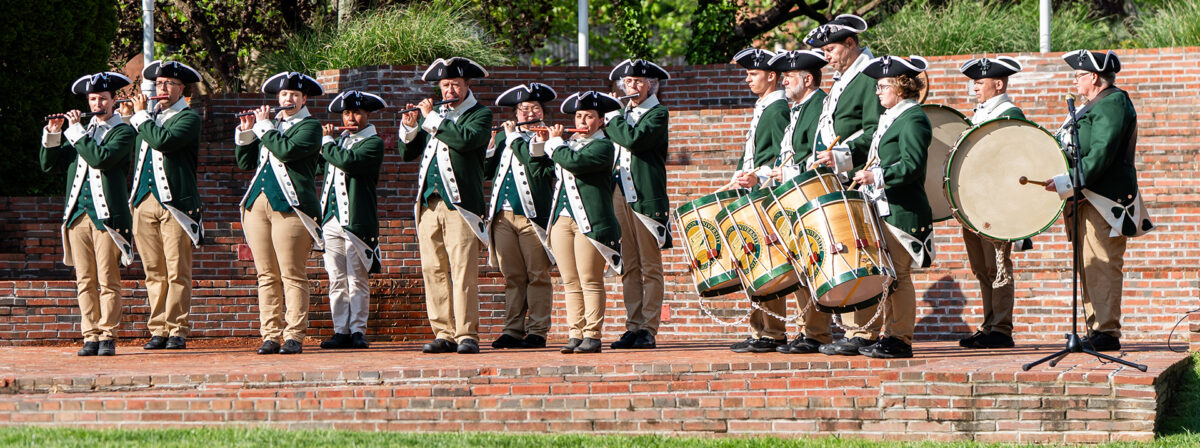 The Patriots Fife and Drum Corps performs on a brick stage in an open air auditorium. It is a sunny day and they are mid-performance. They are wearing green and white colonial era military uniforms with black tricorn hats and khaki pants.