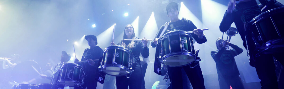 The spirit drumline performs on stage at the anthem in blue-white spotlights with Griz. The picture is close up to the stage looking up at the band.