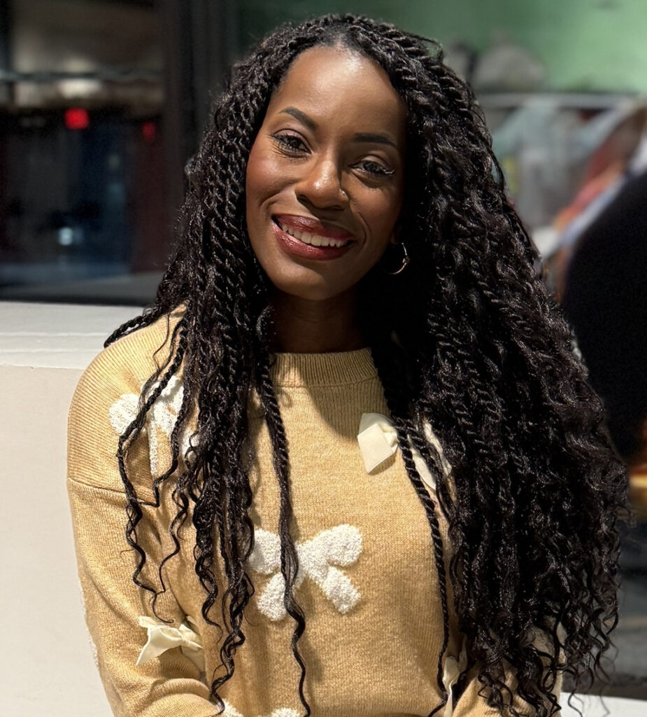 Jennifer is smiling while seated in a blurred theater setting. Jennifer has long black hair. She is wearing a gold sweater with white crocheted bows.