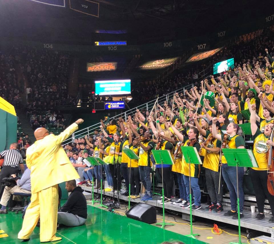 The green machine pep band cheers together with Doc Nix while standing in the stands of EagleBank Arena. Everyone has their right hand in the air. They are wearing gold jerseys and Doc Nix is wearing a yellow suit.