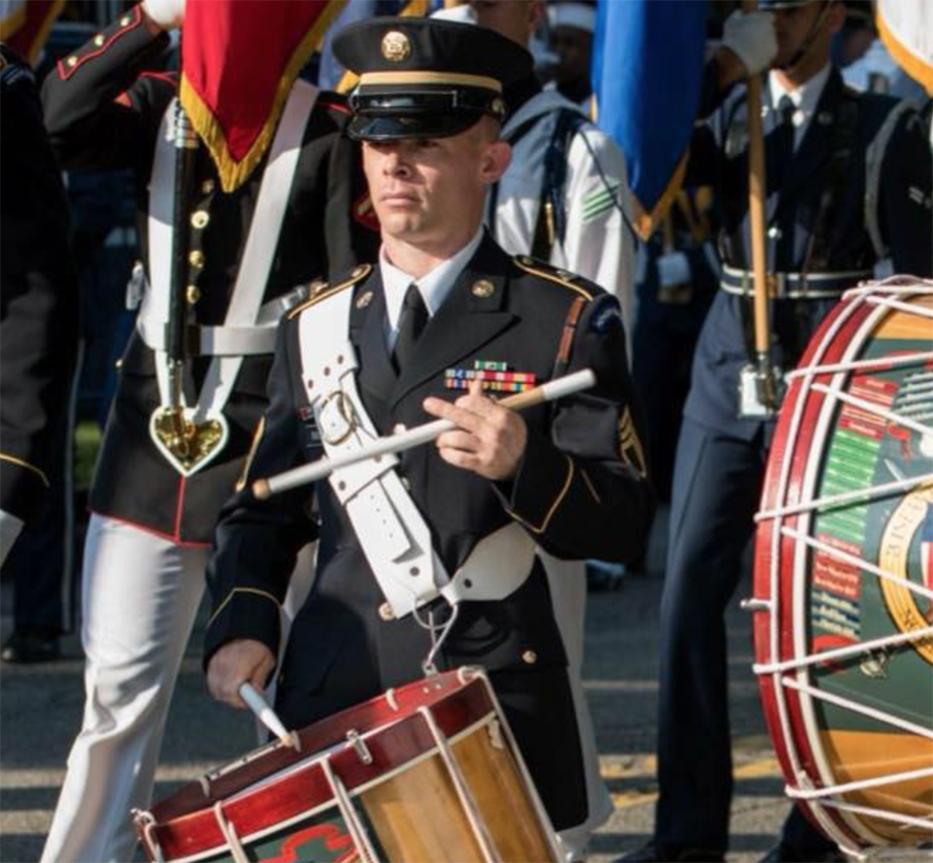 Shane performs on his snare drum with the Army Old Guard Fife and Drum ensemble in his U.S. Army dress uniform during an outdoor procession on a sunny day.