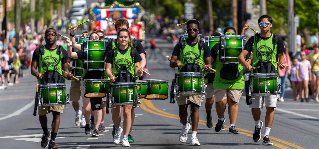 Members of the spirit drumline march down the middle of the street during the Apple Blossom parade in Winchester, Virginia. It is a bright day. They are wearing electric green jerseys with black print and black t shirts and shorts. They are in formation.