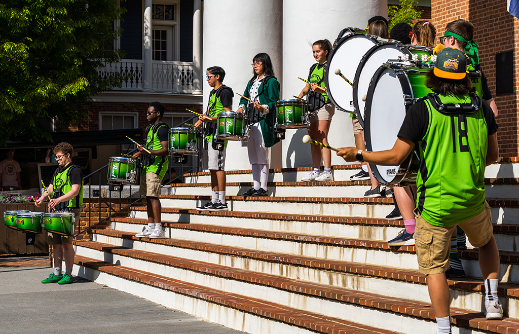 Members of the Green Machine Spirit Drumline stand in a V-formation on steps in front of a Georgian style brick building with large white columns. They are mid-performance in bright green jerseys and shorts.