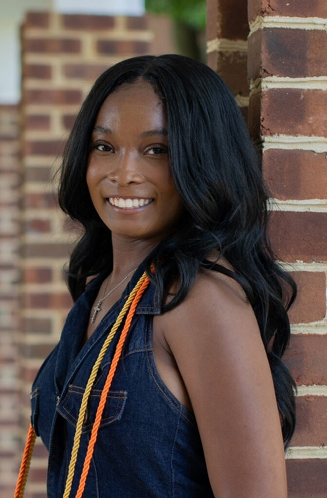 Syla is smiling and standing in front of a brick wall. She has long black hair and is wearing a denim shirt and yellow and orange cords.