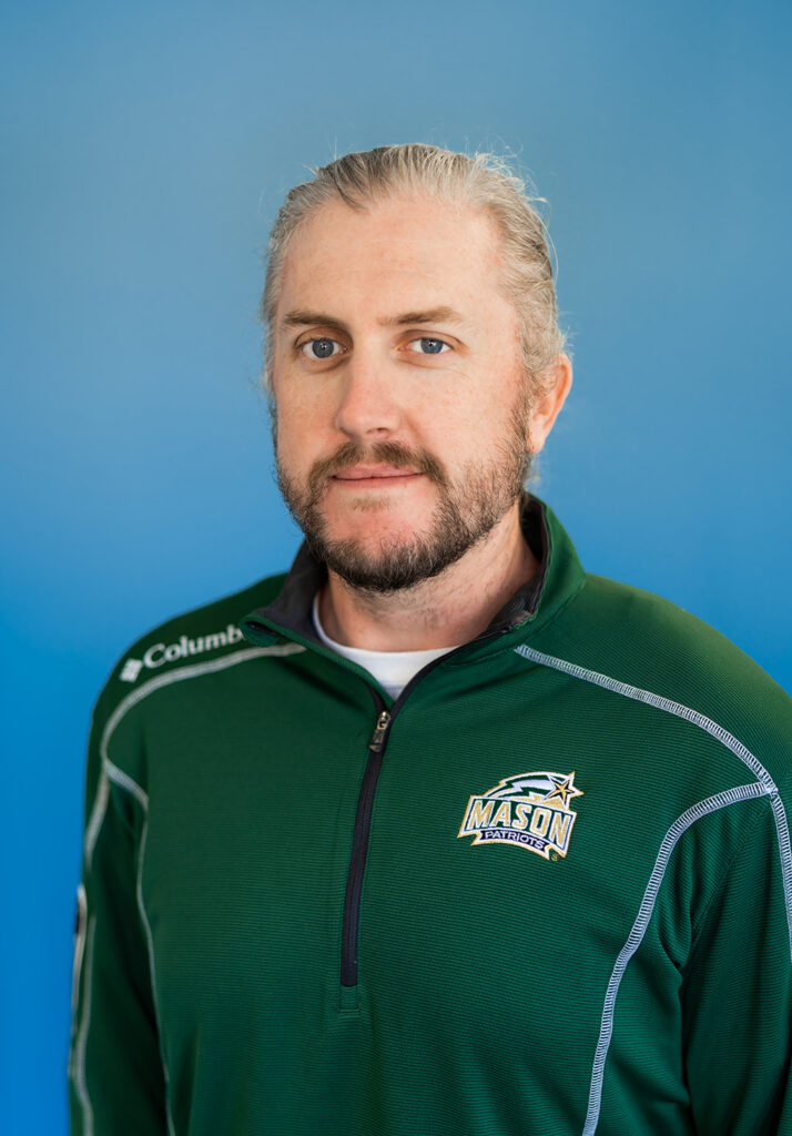 Travis is smiling while seated in front of a blue background. Travis has long blond hair pulled back and a beard. Travis is wearing a green Mason and Columbia branded zip up long sleeve shirt.