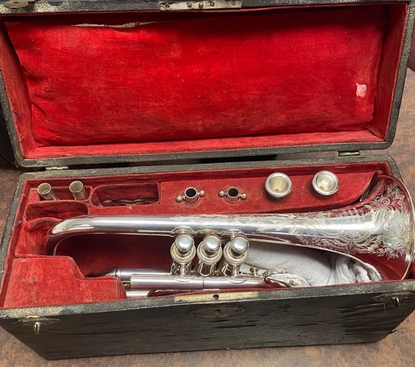 A silver Bb cornet by Besson in an original black case with red lining. The bell is highly engraved with floral patterns. There are 2 mouthpieces, two leadpipes, a tuning slide, and a lyre in the case as well.