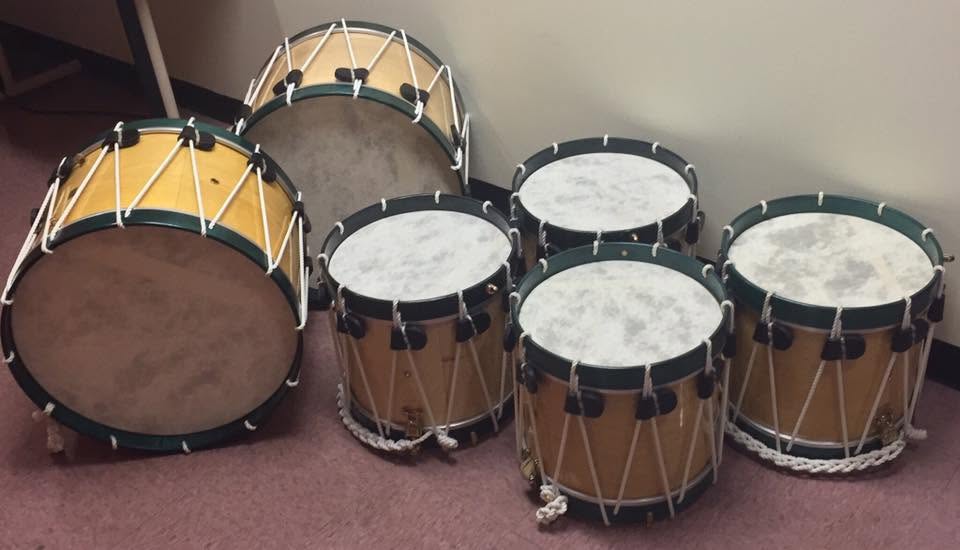 Four snare drums and 2 bass drums, all with a light wood shell, green rims, dark brown leather ears, and white rope
