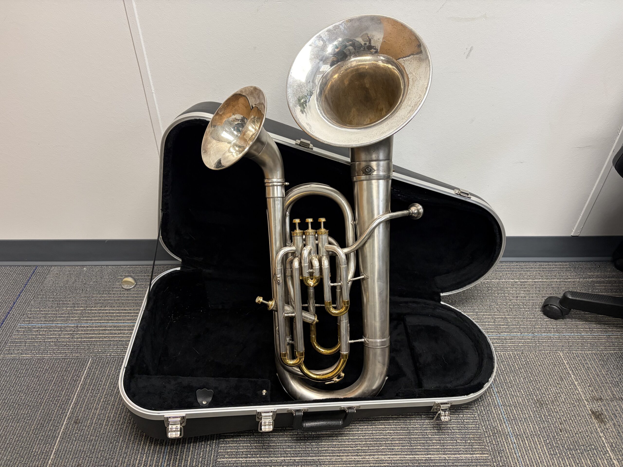 This silver euphonium features two bells both curved outwards. It has three top action piston valves and a fourth piston valve on the side to operate the small bell. The valve caps and slides are gold plated while the rest of the instrument is satin silver. The instruments is standing upright in its black case.