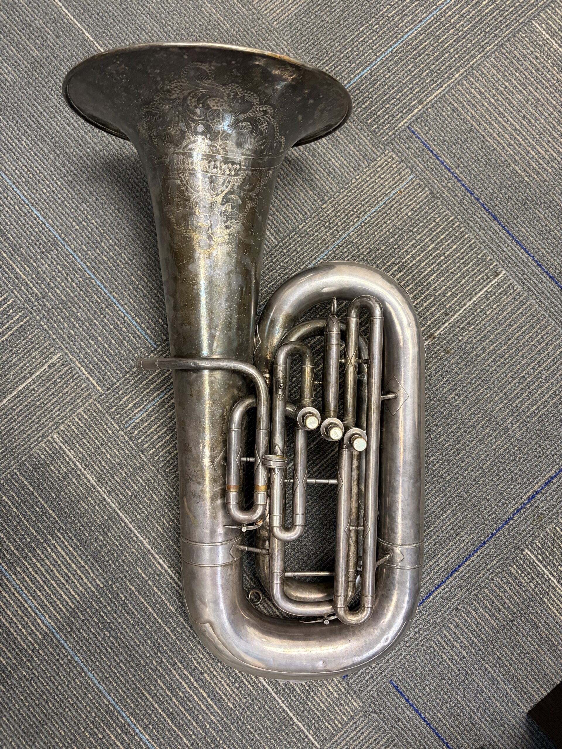A silver plated Eb tuba that reads "Buescher" on the bell and features 3 front-facing piston valves on the side