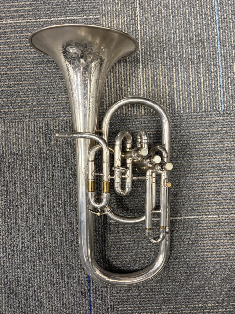 A silver plated Eb alto horn resting on a grey carpet. The instrument features three side action piston valves and a flowery bell engraving.