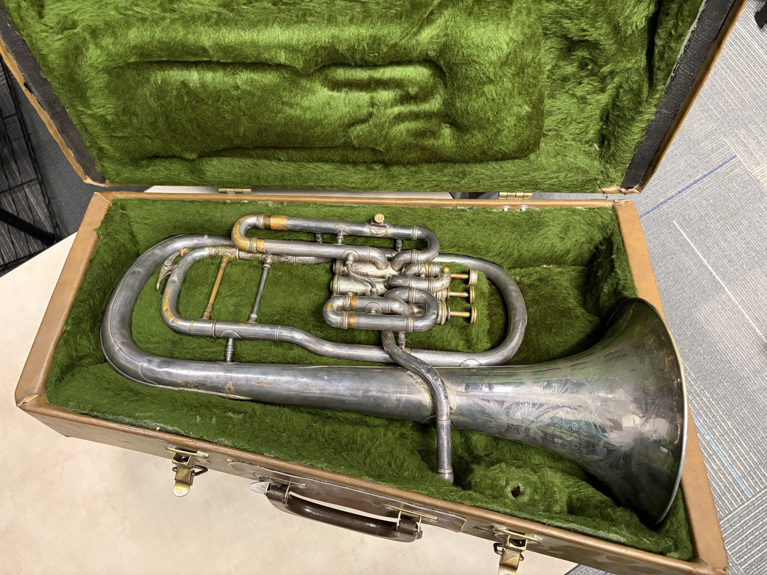 A silver plated Eb alto horn with three upright piston valves. The instrument is laying in its case with a green lining.