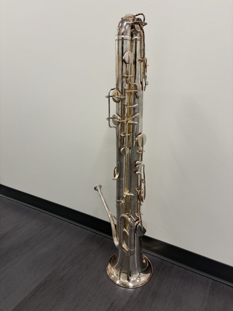 A silver brass instrument standing on its bell on a dark hardwood floor against a white wall. The instrument is approximately 4 feet tall, is silver plated, and features 11-keys.