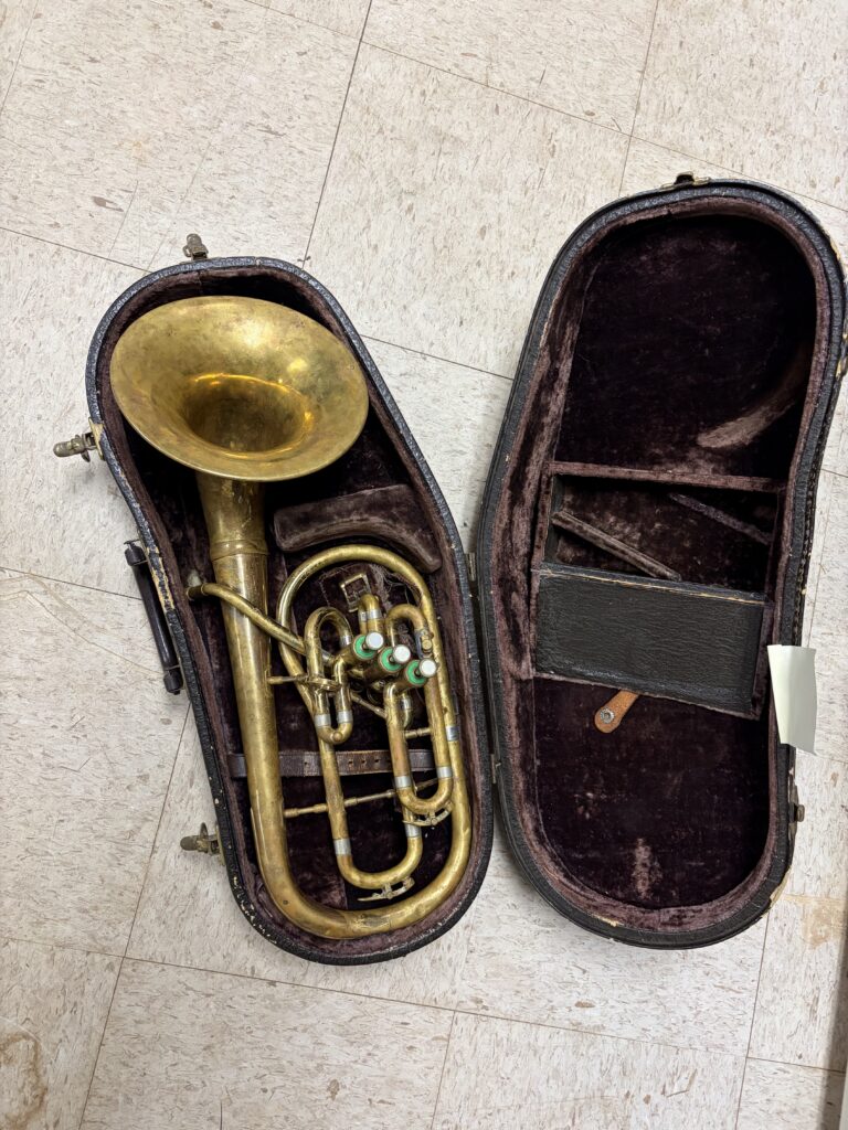 Eb alto horn with bell-front design and 3 side action piston valves. Resting in its case on a dirty white tile floor