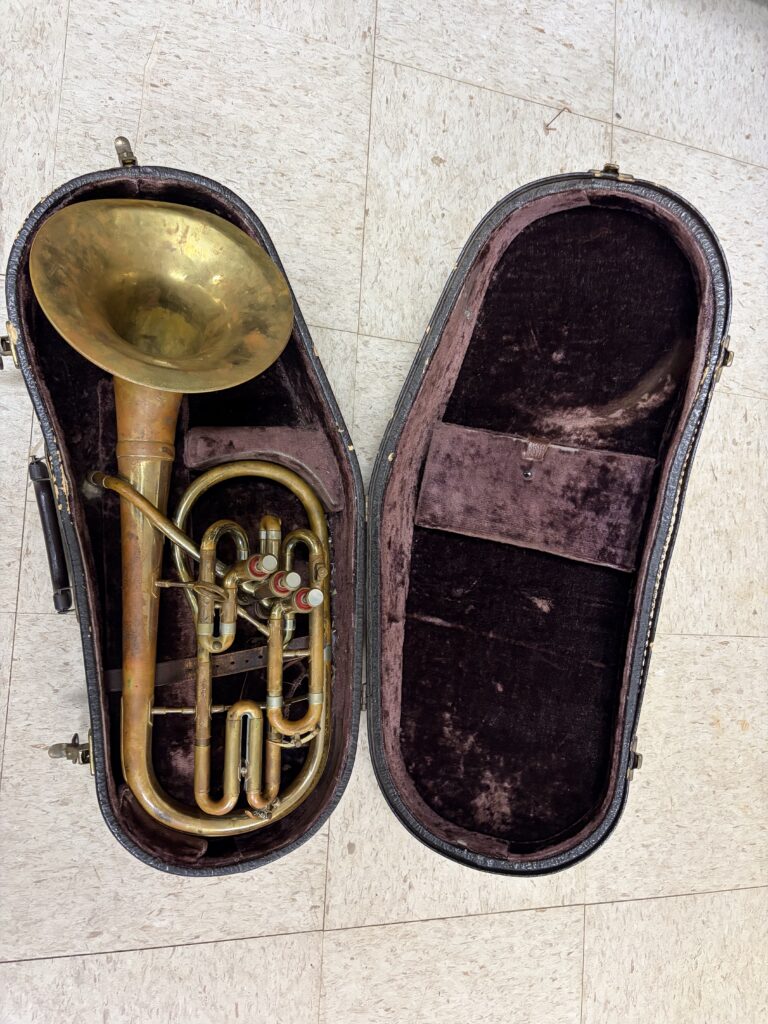 Eb alto horn with bell-front design and 3 side action piston valves. Resting in its case on a dirty white tile floor